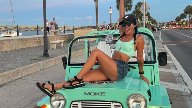 A woman wearing a mint tank top and shorts sits on the hood of a mint-green Moke car by a coastal road.