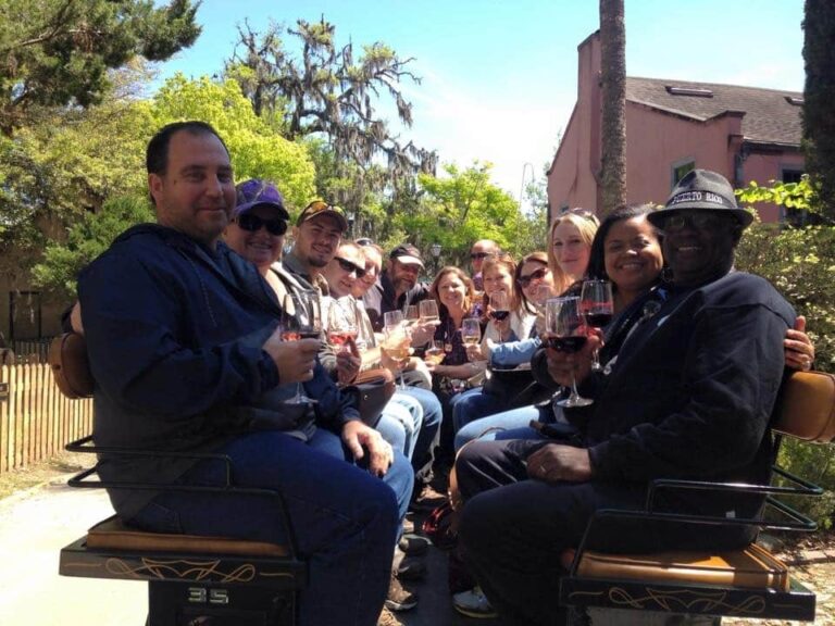 A large, diverse group of friends sits on a mobile patio vehicle outdoors, holding wine glasses and smiling.