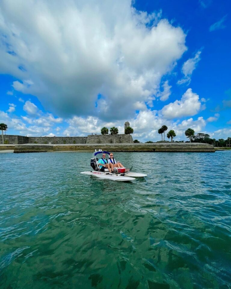 Two people pedal a small, white boat on turquoise water before a stone fort under a bright, cloudy blue sky.