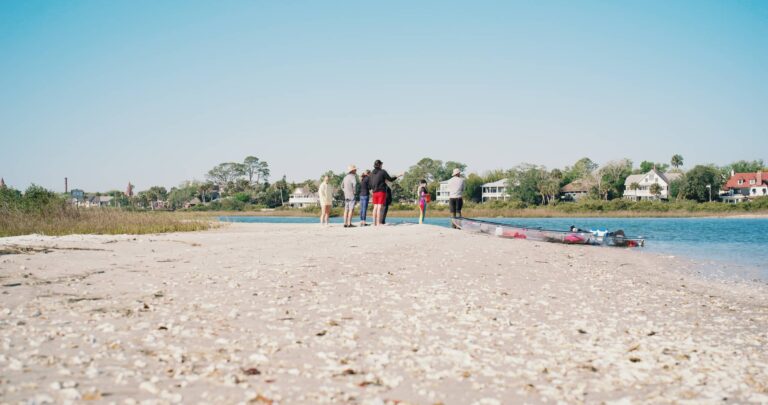 A group of people stand on a sandy, shell-covered shore by a waterway with houses in the background.