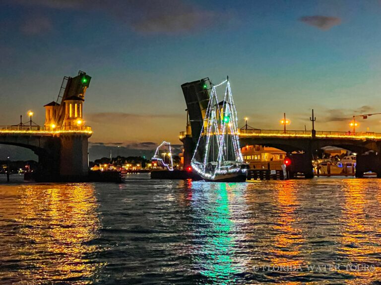 A lit-up sailboat passes through an open drawbridge on the water at dusk, reflecting orange and green lights.